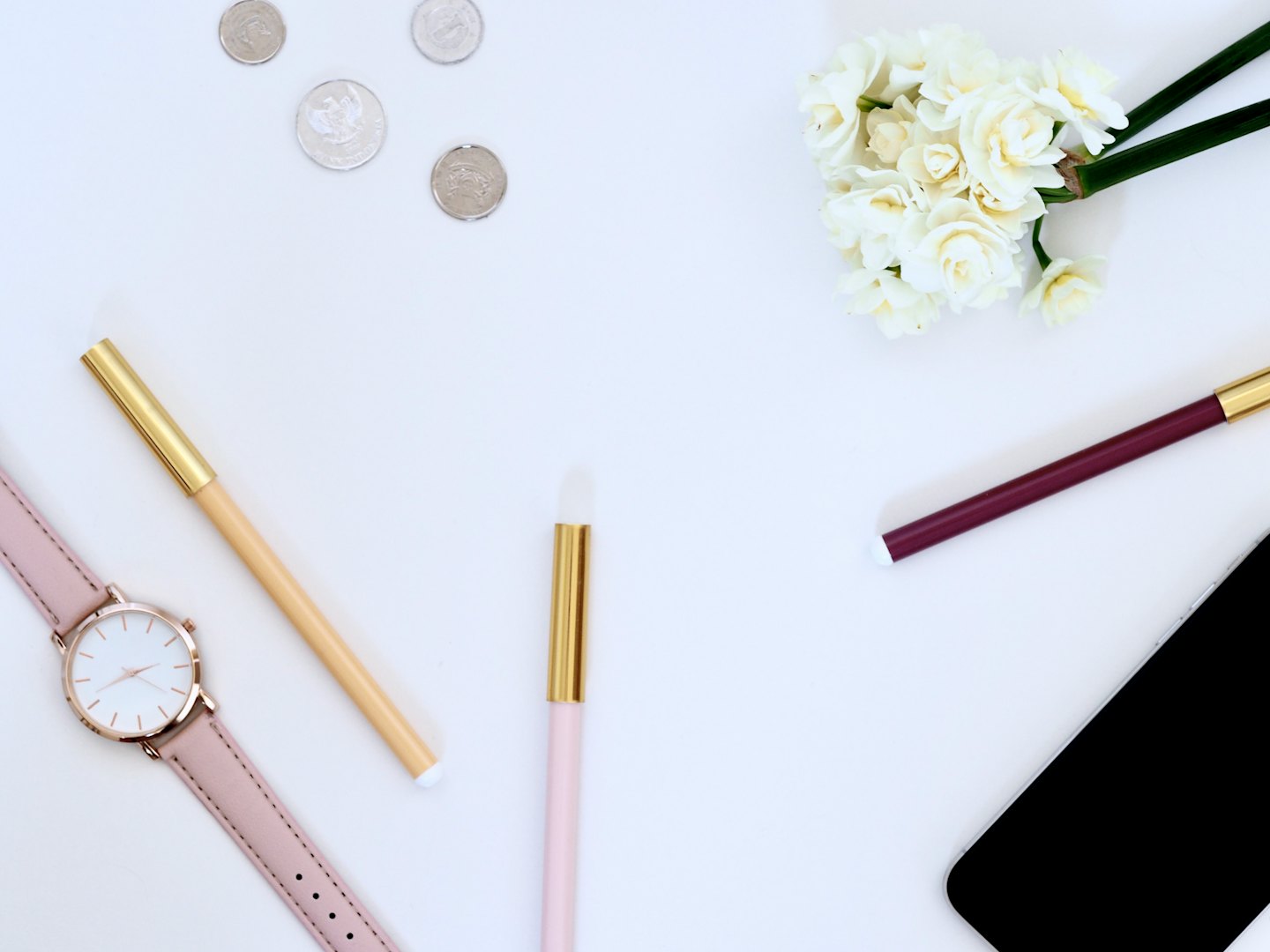 Rose gold watch, pens, coins, narcissus flowers, and smartphone on white surface.