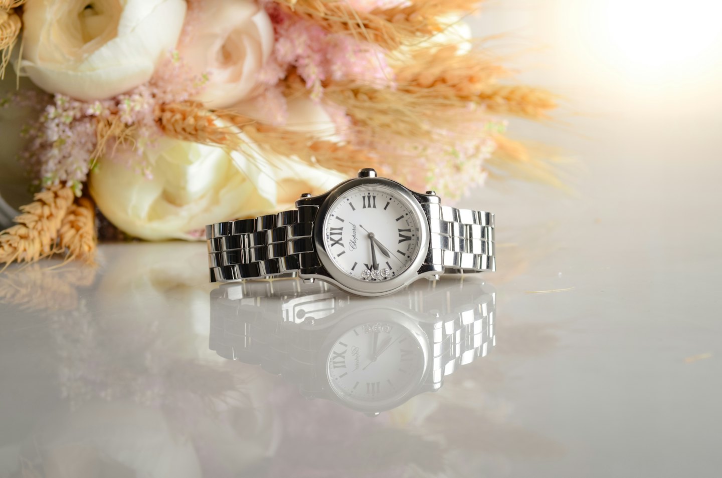 A silver luxury wristwatch with Roman numerals rests on a reflective surface.
